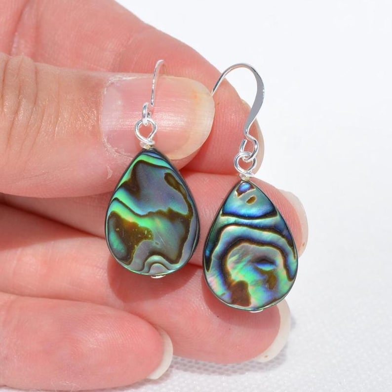 May include: A pair of teardrop-shaped earrings with iridescent abalone shells. The earrings have silver-colored hooks. The abalone shells display a mix of green, blue, and brown colors, with a swirling pattern.