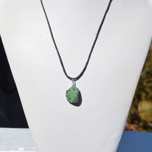 May include: A green gemstone pendant on a black cord necklace. The pendant is a rough, natural stone with a dark green color and white flecks.