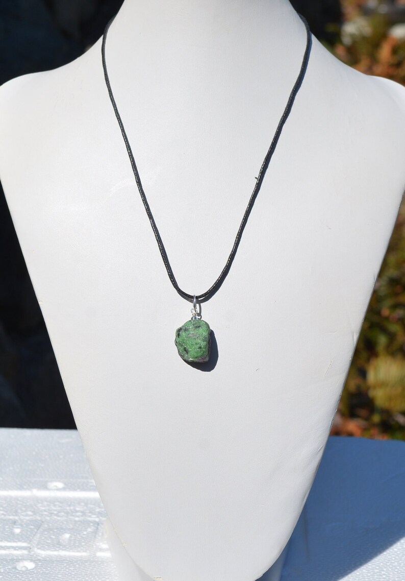 May include: A green gemstone pendant on a black cord necklace. The pendant is a rough, natural stone with a dark green color and white flecks.