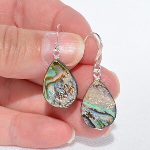 May include: A pair of teardrop-shaped earrings with iridescent abalone shells. The earrings have silver-colored hooks. The abalone shells display a mix of colors, including green, blue, and brown, with a textured surface.