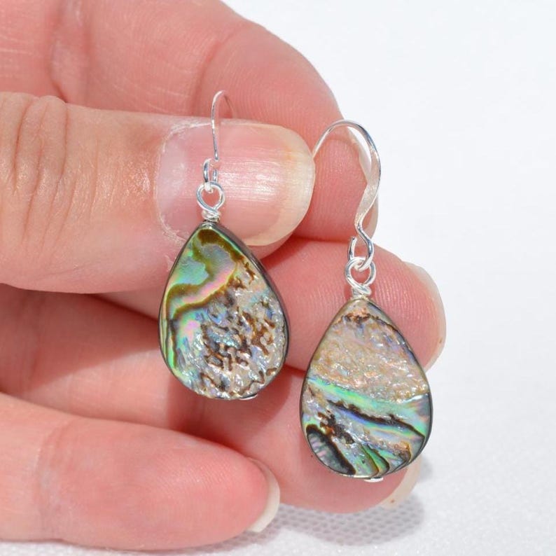 May include: A pair of teardrop-shaped earrings with iridescent abalone shells. The earrings have silver-colored hooks. The abalone shells display a mix of colors, including green, blue, and brown, with a textured surface.