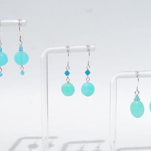 May include: Three pairs of drop earrings featuring round, light turquoise beads suspended from silver-toned hooks. Each earring includes a small, bright blue faceted accent bead above the larger round stone, creating a consistent design across all sets.