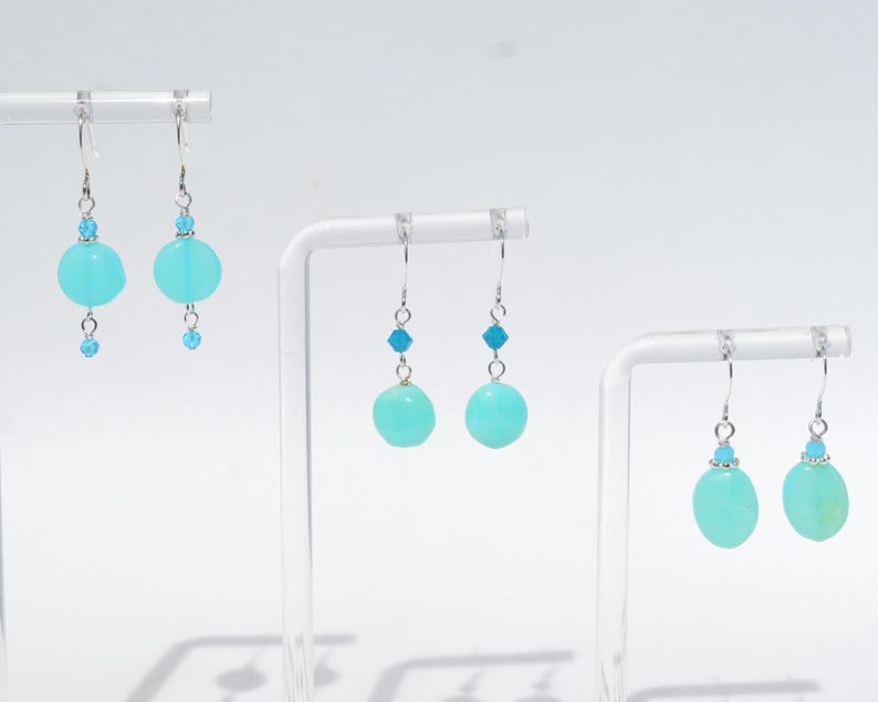 May include: Three pairs of drop earrings featuring round, light turquoise beads suspended from silver-toned hooks. Each earring includes a small, bright blue faceted accent bead above the larger round stone, creating a consistent design across all sets.