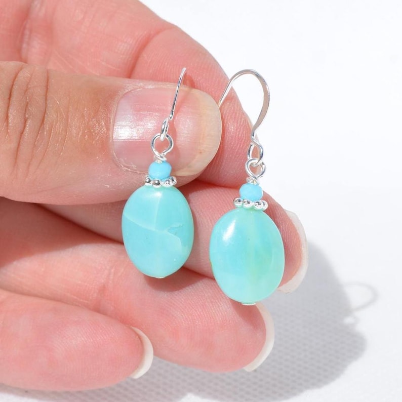 May include: A pair of drop earrings featuring smooth, oval-shaped light blue stones. Each stone is topped with a smaller, round light blue bead and a silver-toned metal finding, suspended from a simple silver-toned curved ear wire hook.