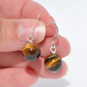 Puede incluir: Un par de pendientes colgantes con piedras preciosas de ojo de tigre redondas y pulidas, con bandas chatoyantes de color marr&oacute;n dorado. Cada cuenta esf&eacute;rica est&aacute; suspendida de un gancho de metal plateado, creando un dise&ntilde;o colgante.