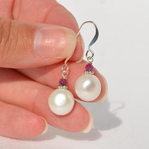 May include: A pair of dangle earrings featuring large, lustrous white pearls. Each earring has a small cluster of red gemstones above the pearl and a silver-colored hook. The earrings are held up by a person's hand.