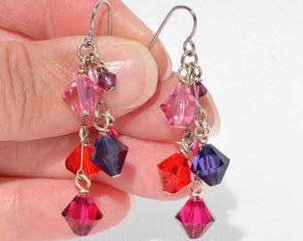 May include: A pair of dangle earrings featuring multiple faceted crystal beads in shades of pink, red, and deep purple. The beads are suspended from silver-toned metal chains and hooks, creating a layered, multi-strand drop design.