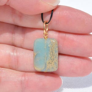 May include: A rectangular pendant with a light blue and tan stone. The stone is suspended from a gold-colored bail and a black cord. The pendant is held by a hand.
