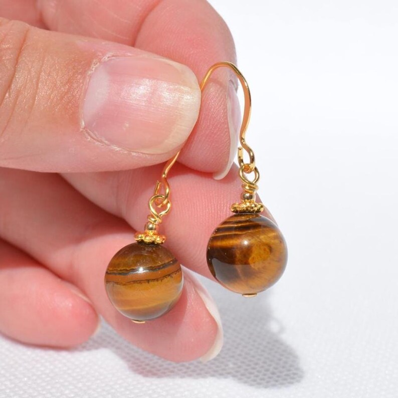 Puede incluir: Un par de pendientes colgantes con piedras preciosas de ojo de tigre redondas y pulidas, con bandas marrones y doradas caracter&iacute;sticas. Cada piedra cuelga de un gancho de metal dorado mediante una peque&ntilde;a cuenta decorativa dorada.
