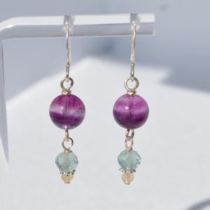 May include: A pair of dangle earrings featuring a large, round, purple and white striped bead. Below, a smaller, faceted, light blue bead is suspended, followed by a tiny, round, yellow bead. The earrings have silver-colored hooks.
