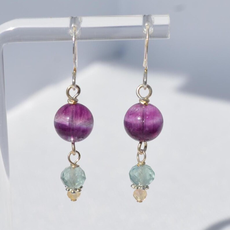 May include: A pair of dangle earrings featuring a large, round, purple and white striped bead. Below, a smaller, faceted, light blue bead is suspended, followed by a tiny, round, yellow bead. The earrings have silver-colored hooks.