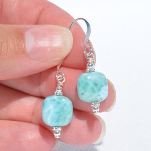 May include: A pair of dangle earrings featuring light blue and white square-shaped beads. The beads are connected to silver-colored hooks with small silver accents. The earrings are held in a hand against a white background.