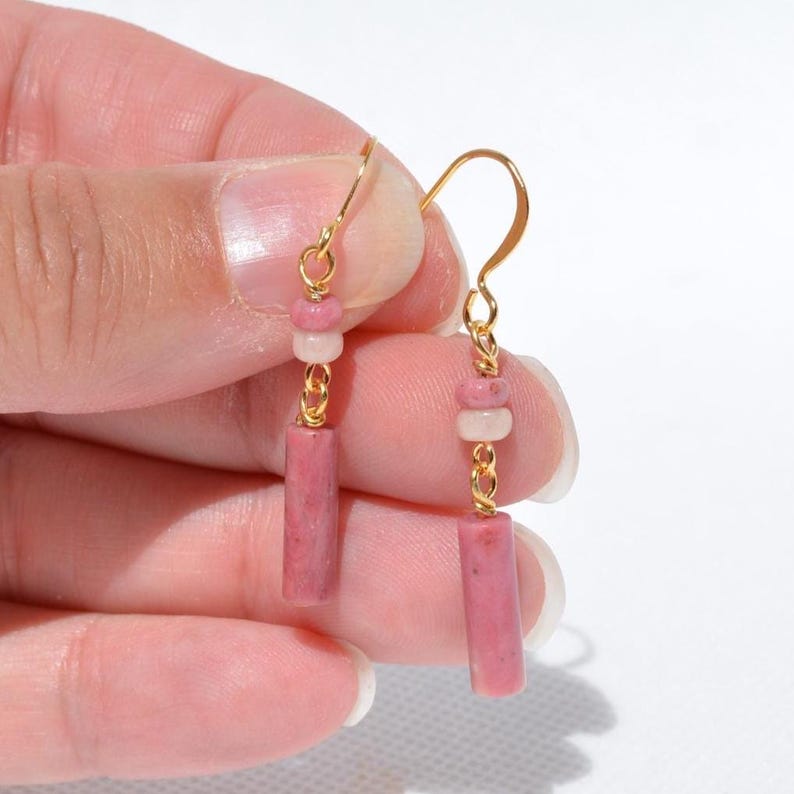 May include: A pair of gold-toned hook earrings featuring a dangling design. Each earring consists of a small round pink bead, a smaller white bead, and a cylindrical pink stone bead suspended from a thin gold-coloured metal wire hook.