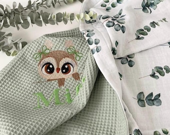 May include: A green waffle weave baby blanket with an embroidered owl and the name "Mia". The blanket is paired with a white muslin blanket with a eucalyptus leaf print.