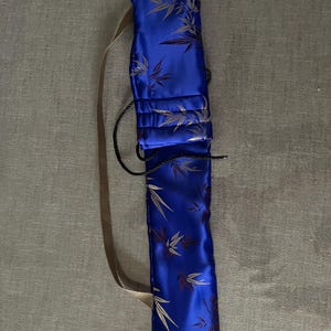 May include: A long, rectangular, royal blue fabric case featuring a repeating pattern of dark brown and gold bamboo leaves. The case includes a tan shoulder strap and a black drawstring closure at the top to secure the contents inside the sleeve.