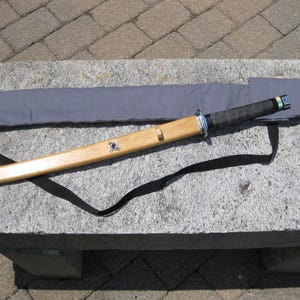 May include: A wooden katana sword with a black handle and a light brown sheath. The sheath is gray and has a black strap. The sword is resting on a gray stone surface.