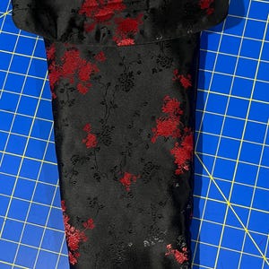 May include: Black satin wine bottle bag with a red floral pattern. The bag has a black strap and a fold-over top. The floral design features red flowers and leaves on a black background. The bag is designed to hold a standard wine bottle.