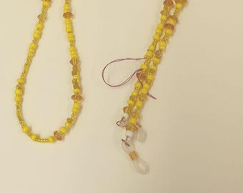 May include: A yellow beaded eyeglass chain with gold accents and clear plastic ends. The chain is made of small round beads and larger, round, amber-colored beads.