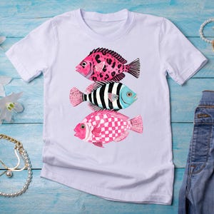 May include: A white short-sleeve t-shirt featuring a graphic print of three stacked fish. The top fish has a pink and black leopard print, the middle fish has black and white vertical stripes, and the bottom fish has a pink and white checkerboard pattern.