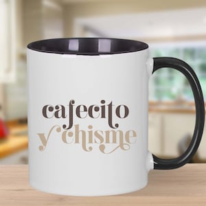 May include: White ceramic coffee mug with a black rim and handle. The mug has the text "cafecito y chisme" printed on it in brown and tan lettering.