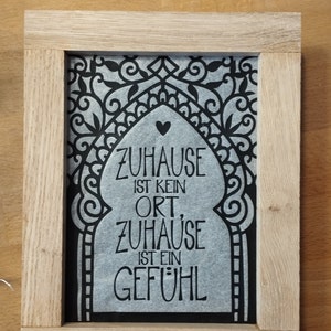 Può includere: Una stampa incorniciata con un design in bianco e nero di un arco stilizzato con un cuore in cima. Il testo "Zuhause ist kein Ort, Zuhause ist ein Gefühl" è stampato in nero su uno sfondo grigio.