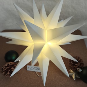 Op de afbeelding: Een witte, verlichte stervormige kerstversiering met meerdere punten. De ster wordt van binnenuit verlicht en geeft een warme gloed. Het woord "Christmas" staat op een klein label. Decoratieve dennenappels en ornamenten omringen de ster.
