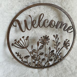 Könnte beinhalten: Ein rundes Holzschild mit dem ausgeschnittenen Wort "Welcome" und einem floralen Design am Rand.