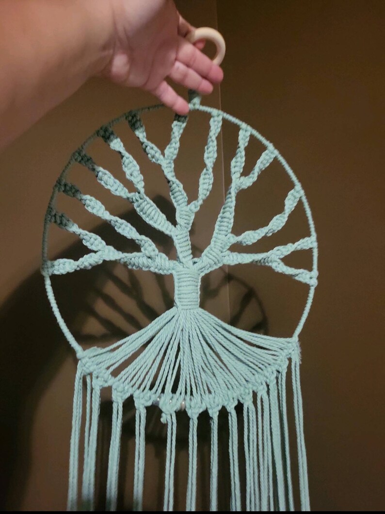 May include: A handmade macrame wall hanging featuring a tree design in a circular frame. The tree is crafted with light teal-colored cord, with a wooden ring at the top for hanging. The design includes a detailed tree trunk and branches.
