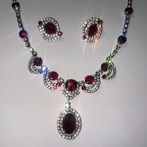 May include: A jewelry set consisting of a necklace and a pair of matching earrings. The necklace features a central oval red gemstone pendant surrounded by clear crystals, with additional red stones and clear crystal accents along the silver-tone chain.