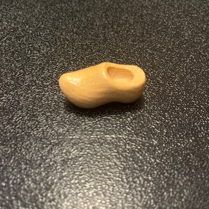 May include: A small, wooden clog in a light tan color. The miniature shoe has a hollowed-out interior and is resting on a dark, textured surface. This decorative item is a charming representation of traditional Dutch footwear.