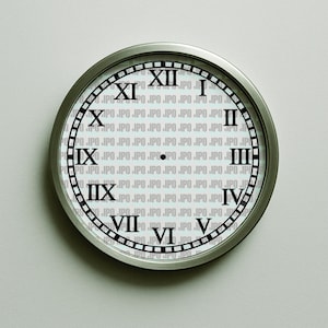 May include: A round wall clock with a silver-colored metal frame. The clock face is white with black Roman numerals and a black minute and hour hand. The background of the clock face has a repeating pattern of the letters "IPO".