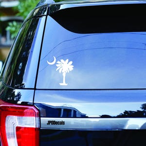 May include: White vinyl decal of the South Carolina state flag, featuring a crescent moon and a palmetto tree. The decal is affixed to the rear window of a black vehicle. The text "JPODecals" is visible.
