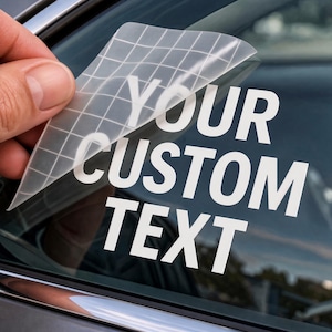 May include: A white vinyl decal with the text YOUR CUSTOM TEXT applied to a car window. The decal features a clear transfer tape with a grid pattern partially peeled back from the surface, revealing the bold, sans-serif lettering underneath.