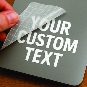 May include: A gray surface with the words "YOUR CUSTOM TEXT" in white, with a hand peeling back a transparent grid-patterned transfer sheet. The image highlights the application of custom text decals.