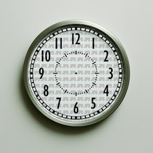 May include: A round, silver-framed wall clock with a white face. Black numerals mark the hours, and black lines indicate the minutes. The clock's face has a subtle, repeating pattern in the background. The clock is mounted on a light gray wall.