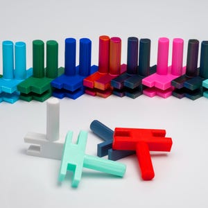 May include: A row of 15 colorful plastic building blocks in various shades of green, blue, red, pink, and purple. The blocks are arranged in a line and have a simple design with two vertical posts and a horizontal connecting piece. There are also four additional blocks in the foreground, including a white block with a vertical post and two horizontal connecting pieces, a turquoise block with a vertical post and two horizontal connecting pieces, a red block with a vertical post and two horizontal connecting pieces, and a blue block with a vertical post and two horizontal connecting pieces.