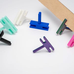 May include: Assortment of colourful plastic shelf brackets in various colours, including white, blue, green, black, purple, and pink. The brackets are designed to hold shelves in place.