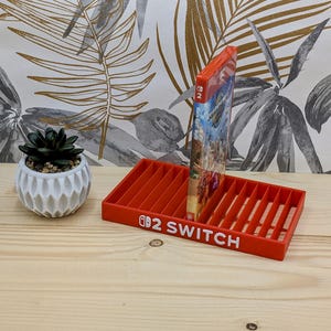 以下が含まれることがあります： 白い文字で「Switch」と書かれた赤いプラスチック製のNintendo Switchゲームホルダー。ゲームカートリッジがホルダーに配置されています。ホルダーの左側には、白い鉢に入った小さな多肉植物があります。