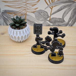 May include: Three black and white 3D printed figurines of the video game character Mr. Game & Watch. The figurines are standing on yellow and black bases. The character on the left is holding a hammer, the character in the middle is holding a bell, and the character on the right is holding a number 8.