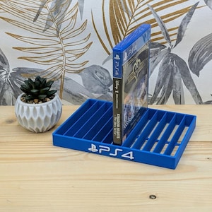 May include: A blue plastic game storage rack with multiple slots for game cases, featuring a white PS4 logo on the front edge. A single blue PS4 game case titled Kingdom Hearts The Story So Far is inserted into one of the slots on the storage rack.