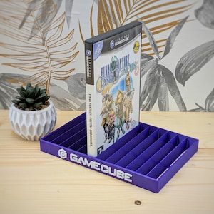 May include: Purple GameCube game case holder with a GameCube logo and the words "GameCube" printed on the front. A purple GameCube game case with the text "Final Fantasy Crystal Chronicles" is standing in the holder.