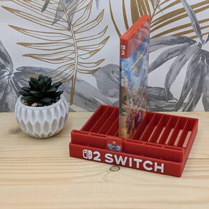 Può includere: Un supporto in plastica rossa per videogiochi con molteplici fessure verticali per le custodie dei giochi e fessure frontali più piccole per le cartucce. La parte anteriore mostra la scritta bianca 2 SWITCH con un logo Nintendo Switch.