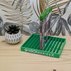以下が含まれることがあります： 白い「XBOX SERIES X」の文字が入った緑色のXbox Series Xゲームホルダー。「FIFA 21」のビデオゲームケースがホルダーに入っています。左側には、白い鉢に入った小さな多肉植物があります。