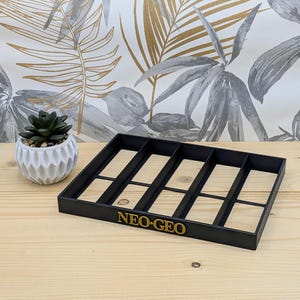Peut inclure: Un plateau de rangement rectangulaire noir en plastique doté de cinq compartiments verticaux et du texte doré NEO-GEO imprimé sur le bord avant. Le plateau présente une base plate et une conception ouverte sur le dessus pour le rangement.