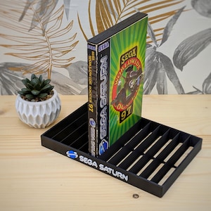 Puede incluir: Un soporte de cartuchos de juego Sega Saturn de plástico negro con un logotipo de Sega Saturn en la parte delantera. El soporte tiene 8 ranuras para cartuchos de juego. Un cartucho de juego Sega Saturn está en el soporte. El cartucho de juego es verde y negro con el texto "Sega Worldwide Soccer 97" en el lateral.