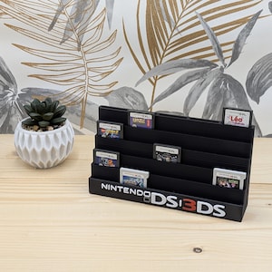 Könnte beinhalten: Ein schwarzer Spielkassettenhalter für Nintendo DS- und 3DS-Spiele. Der Halter hat mehrere Ebenen zur Anzeige von Spielkassetten. Der Text "NINTENDO DS 3DS" ist weiß auf der Vorderseite aufgedruckt. Links eine kleine Sukkulente in einem weißen Topf.