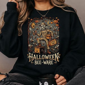 May include: A black long-sleeve sweatshirt featuring a graphic print of a skeleton dressed as a beekeeper, surrounded by bees, pumpkins, and a beehive. The text "HALLOWEEN BEE-WARE" is printed in a cream-colored font below the central illustration.