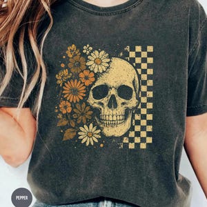 May include: Dark gray t-shirt featuring a skull design with flowers and a checkered pattern. The skull is yellow, and the flowers are in shades of yellow and orange. The shirt has short sleeves and a crew neck.