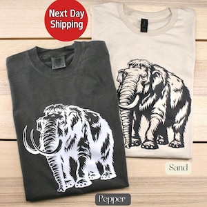 May include: Two t-shirts with a white woolly mammoth graphic. The shirt on the left is dark gray and says "Pepper" below the graphic. The shirt on the right is light beige and says "Sand" below the graphic.