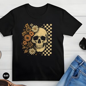 May include: Black t-shirt featuring a skull design with floral accents and a checkered pattern. The skull is cream-colored, and the flowers are in shades of orange and yellow. The shirt is paired with jeans and sunglasses.